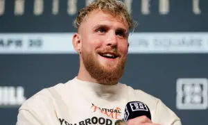 Jake Paul, znany z kontrowersyjnych wypowiedzi i walk, uśmiecha się do mikrofonu. Jego wzrost jest imponujący, co widać po jego postawie.