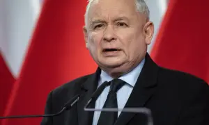 Jarosław Kaczyński, polityk, przemawia na tle flagi Polski. Jego wzrost jest typowy dla mężczyzny w jego wieku.