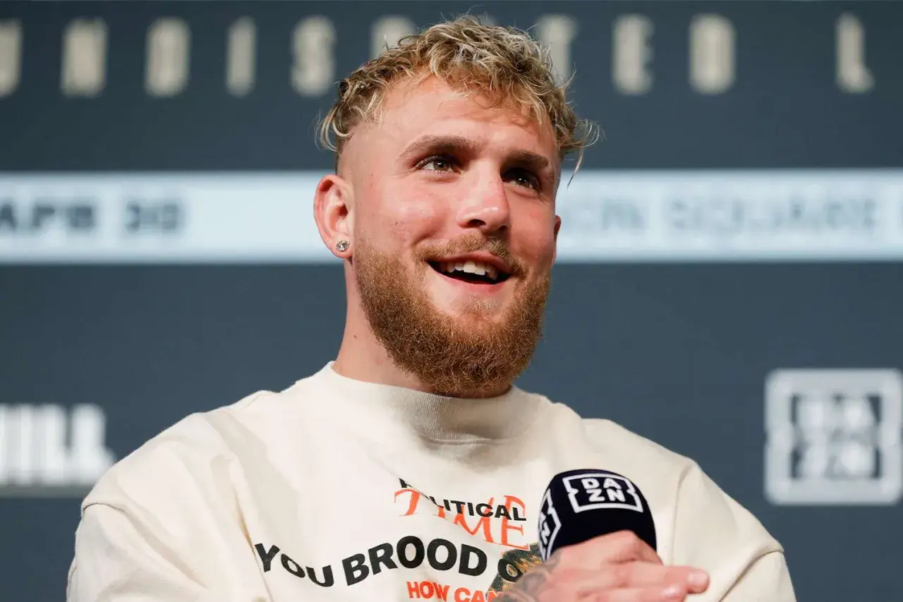 Jake Paul, znany z kontrowersyjnych wypowiedzi i walk, uśmiecha się do mikrofonu. Jego wzrost jest imponujący, co widać po jego postawie.