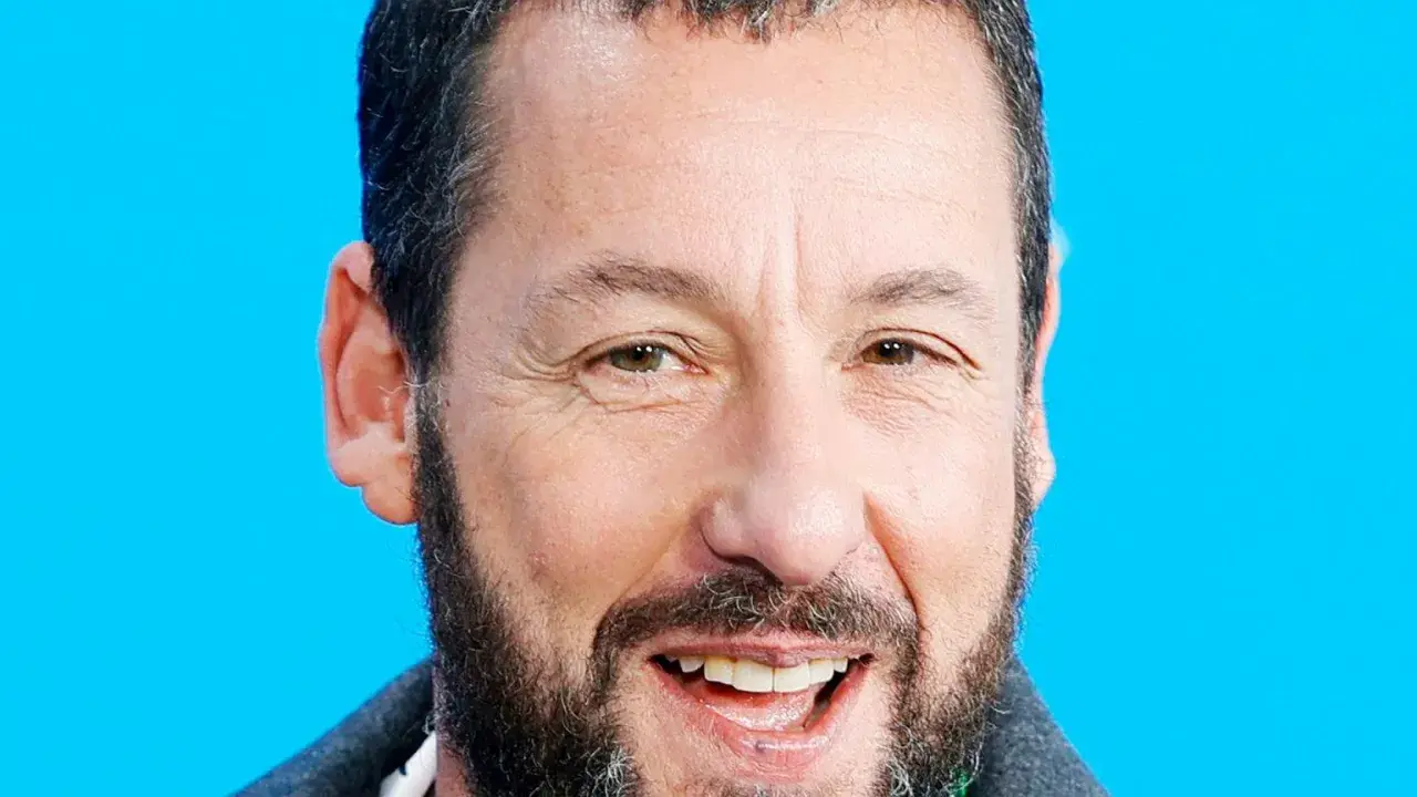 Uśmiechnięty Adam Sandler, gwiazda wielu komediowych filmów, na tle niebieskiego tła.