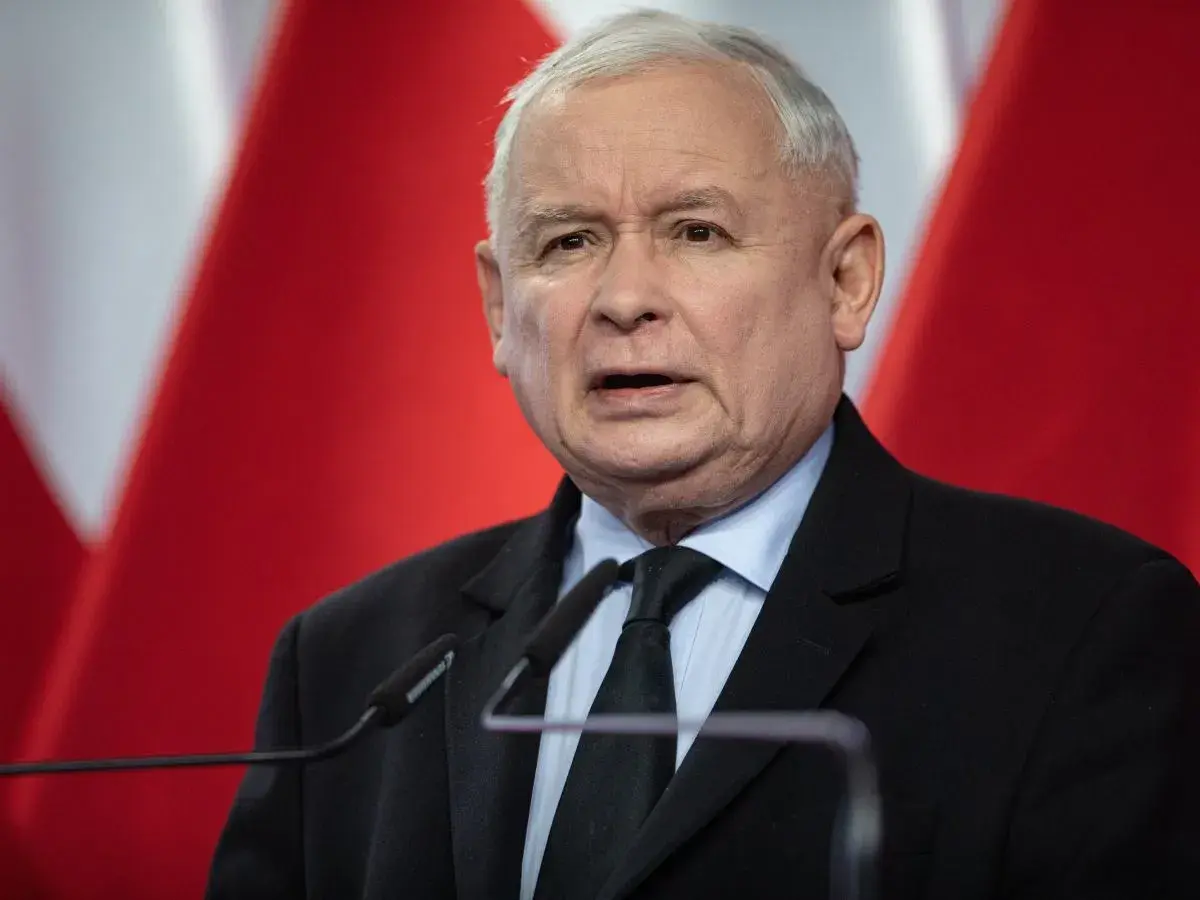 Jarosław Kaczyński, polityk, przemawia na tle flagi Polski. Jego wzrost jest typowy dla mężczyzny w jego wieku.