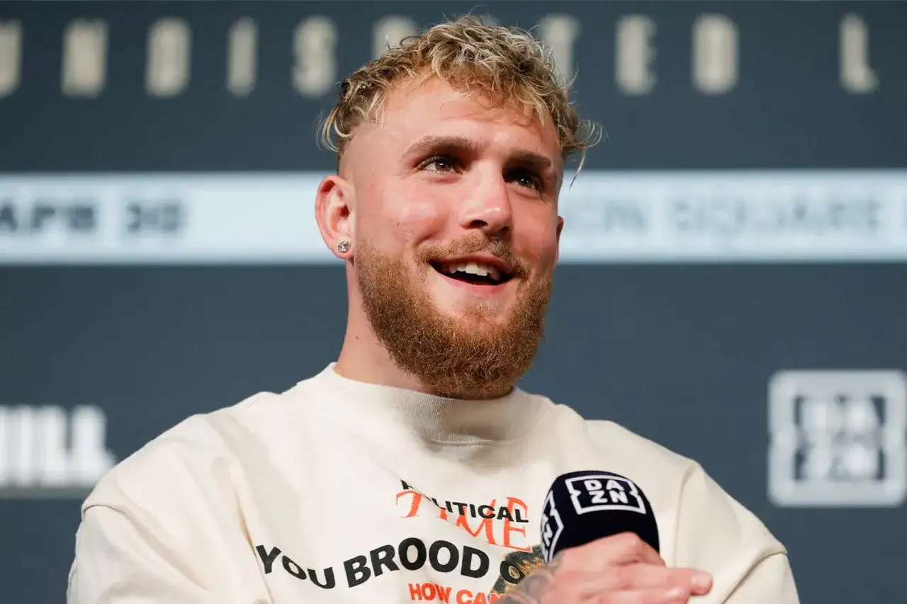 Jake Paul, znany z kontrowersyjnych wypowiedzi i walk, uśmiecha się do mikrofonu. Jego wzrost jest imponujący, co widać po jego postawie.
