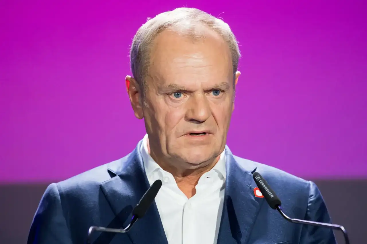 Donald Tusk, polityk o charakterystycznym wyglądzie, przemawia na tle fioletowej sceny. Jego wzrost jest imponujący, a pewność siebie emanuje z każdego gestu.