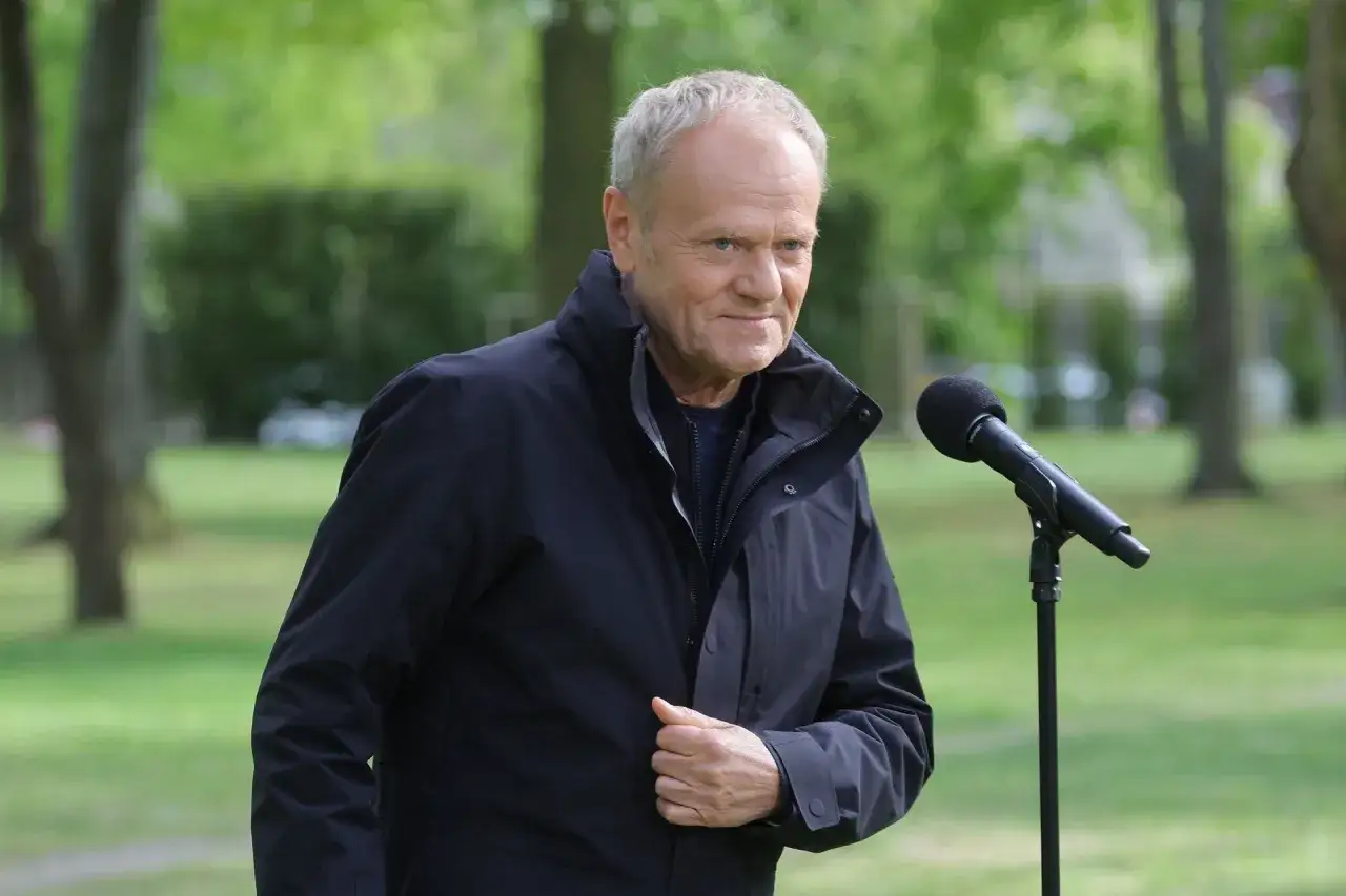 Donald Tusk, w czarnej kurtce, przemawia do mikrofonu. Jego wzrost jest typowy dla mężczyzny w średnim wieku.