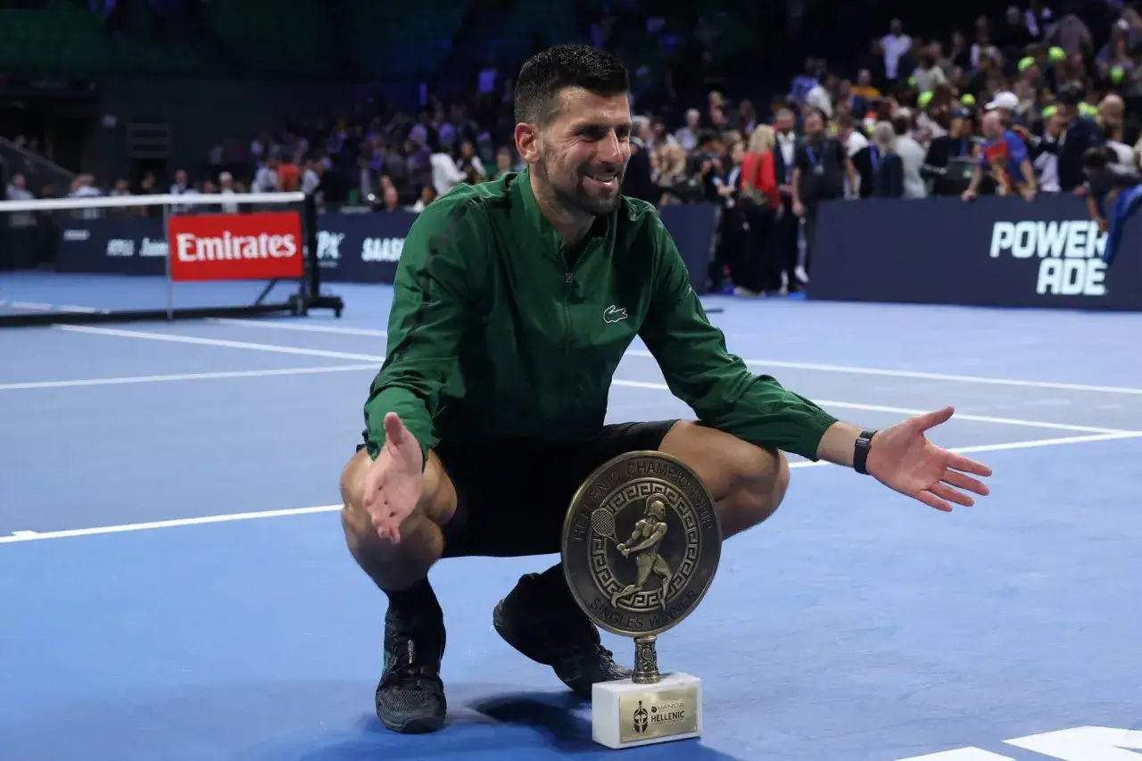 Novak Djokovic, mimo swojego wieku, triumfuje z pucharem. Uśmiechnięty tenisista w zielonej kurtce klęczy na korcie.